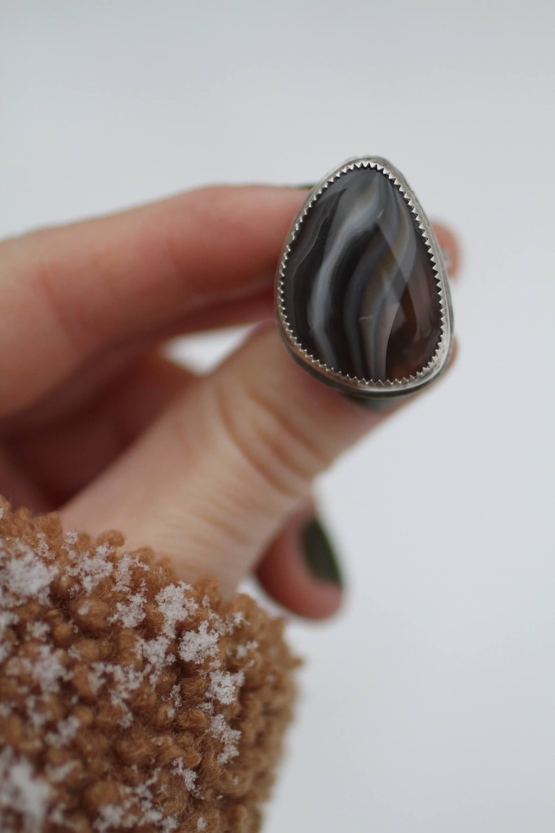 May include: A silver ring with a teardrop-shaped stone. The stone features a pattern of brown, gray, and white bands. The ring has a serrated edge around the stone.