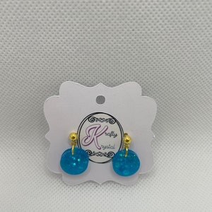 May include: A pair of blue glitter stud earrings with gold posts. The earrings are on a white card with the text "Krafty Crystal" in gold lettering.