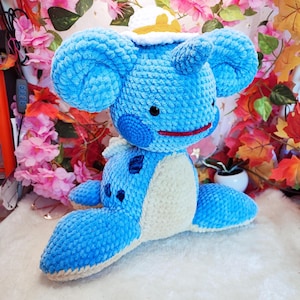 Puede incluir: Un peluche azul hecho a mano con orejas grandes, una cara redonda y una barriga blanca. El juguete tiene ojos negros y una sonrisa roja. Está sobre una superficie blanca, con flores rosas y hojas de otoño en el fondo.