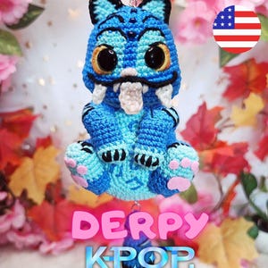 Pode incluir: Um chaveiro de crochê azul feito à mão com um demônio de desenho animado com olhos grandes, presas e uma língua saliente. O chaveiro tem o texto "DERPY KPOP DEMON HUNTERS" em uma fonte colorida e estilizada. O fundo inclui decorações florais e de folhas de outono.