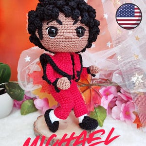 Puede incluir: Muñeca de ganchillo hecha a mano de Michael Jackson con un atuendo rojo con detalles en negro, zapatos negros y un peinado rizado negro. La muñeca está en una postura dinámica de carrera, con el nombre "MICHAEL Jackson" escrito en rojo.