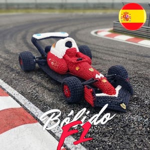 Puede incluir: Un coche de carreras de Fórmula 1 tejido a ganchillo en rojo, blanco y azul oscuro. El coche presenta el número 44 en el morro, un alerón trasero y cuatro ruedas. El texto Bólido F1 y Zoecustomcrafts aparece junto a una bandera española.