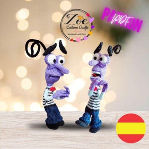 May include: Two crocheted purple and blue plush toys of the character Fear from the movie Inside Out. The toys are standing on a wooden surface. The toys are handmade and have a striped shirt and blue trousers.