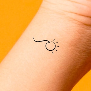 May include: A simple black line drawing of a sun with rays on a person's wrist.