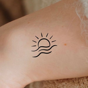 May include: A simple line drawing tattoo of a sun setting over the ocean. The sun is a circle with rays coming out of it, and the ocean is represented by three wavy lines.