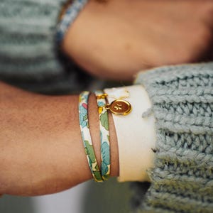 May include: Close-up of two floral print bracelets with gold accents. One bracelet features a gold charm. The bracelets are set against a backdrop of a light green sweater and a white shirt sleeve. Jewelry.
