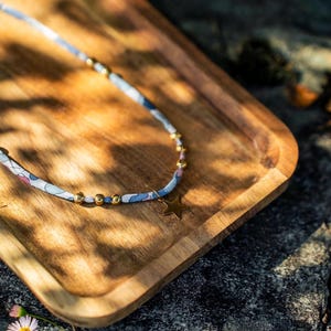 Peut inclure: Un collier délicat avec un cordon en tissu à motifs floraux, orné de perles dorées et d'un pendentif en forme d'étoile. Le collier est posé sur un plateau en bois, avec le mot "weco" en lettres dorées. L'ensemble est éclairé par la lumière naturelle.