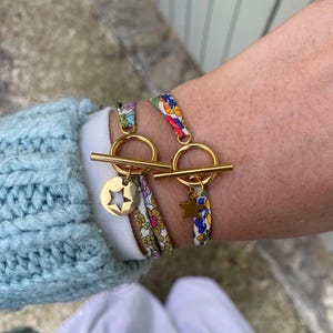 Könnte beinhalten: Zwei goldfarbene Armbänder mit floralen Stoffbändern und Knebelverschlüssen. Eines hat einen goldenen Sternanhänger, das andere eine goldene Scheibe mit Sternaussparung. Die Armbänder werden am Handgelenk getragen.