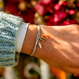 Può includere: Un braccialetto d'argento con un ciondolo dorato e dettagli di perle. Il braccialetto è indossato su un polso, con un maglione azzurro e una camicia bianca visibili sullo sfondo. Il ciondolo è una conchiglia dorata con un piccolo accento arancione.