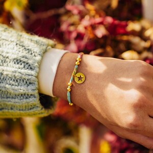 Peut inclure: Un bracelet de perles avec un charm en forme d'étoile dorée. Le bracelet est composé de perles turquoise, dorées et roses. Le charm comporte une étoile noire au centre. Le bracelet est porté sur un poignet avec un pull bleu clair et une chemise blanche.