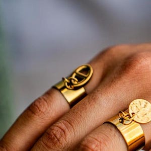 Può includere: Primo piano di due anelli d'oro. Uno presenta una fascia rettangolare con un elemento decorativo, mentre l'altro ha un ciondolo circolare. Gli anelli sono indossati alle dita, mostrando il loro design e la lucentezza metallica. Lo sfondo è sfocato.