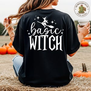 May include: Black t-shirt with the words "basic WITCH" in white script and a witch on a broom graphic. The shirt is a relaxed fit and suitable for Halloween or everyday wear.