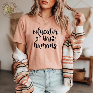 Op de afbeelding: Een perzikkleurig T-shirt met de tekst "educator of tiny humans" in zwart schrift. Het shirt wordt gecombineerd met een lichtblauwe spijkerbroek en een gestreepte trui. Een logo met een cactus is linksboven zichtbaar.