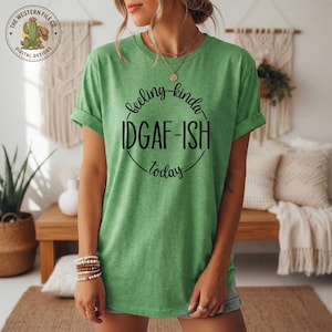 Op de afbeelding: Groen T-shirt met opgerolde mouwen met de tekst "feeling kinda IDGAF-ISH today" in een zwart cirkelvormig ontwerp. Het shirt heeft een ronde hals en is gemaakt van een zacht materiaal. Het logo van The Western File Co. is linksboven zichtbaar.