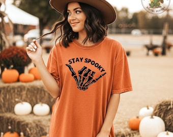 May include: An orange t-shirt with the words "Stay Spooky" and a black skeleton hand graphic. The person in the image is wearing a brown hat. Pumpkins and hay bales are in the background, suggesting a fall or Halloween theme.
