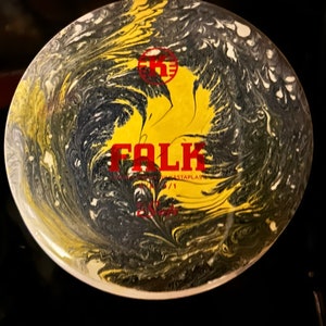 May include: A yellow and black disc with a swirling pattern. The disc has the word "FOLK" printed on it in red letters. The disc is made by "Kastaplast".
