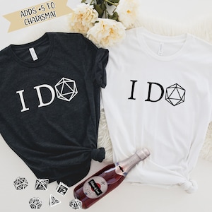 May include: Two t-shirts with the text "I DO" and a black and white image of a twenty-sided die. The shirts are black and white. A bottle of rose wine and several dice are on a white surface.