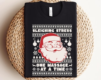 May include: Black t-shirt with a white Christmas-themed design featuring Santa Claus and the text "Sleighing Stress One Massage At A Time".