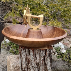 Könnte beinhalten: Eine handgefertigte Holzschale mit einem Nussknackermechanismus aus Messing. Die Schale ist aus dunklem Holz gefertigt und steht auf einem Baumstumpf. Der Nussknacker hat ein Schiffsraddesign. Die Schale hat einen Durchmesser von ca. 30 cm.
