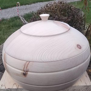May include: A round wooden bowl with a lid, made of light wood with a natural finish. The bowl has a smooth, rounded shape and is made of several pieces of wood that have been joined together.