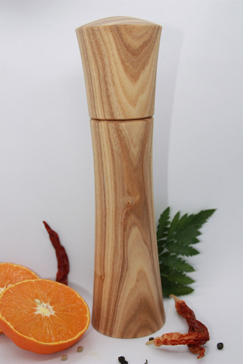 K&ouml;nnte beinhalten: Eine Pfefferm&uuml;hle aus Holz mit einer nat&uuml;rlichen Holzmaserung. Die M&uuml;hle ist hoch und schlank mit einem abgerundeten Kopf. Sie steht auf einer wei&szlig;en Oberfl&auml;che mit Orangenscheiben, roten Chilischoten und schwarzen Pfefferk&ouml;rnern.