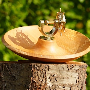 May include: A wooden bowl with a brass ship's wheel nutcracker. The bowl is light brown and sits atop a tree stump. The nutcracker is gold-colored and has a detailed ship's wheel design. The background is a blurred green.