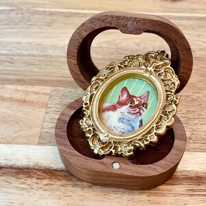May include: A small, ornate gold frame with a painted portrait of a white and brown cat inside. The frame is sitting in a wooden box with a velvet lining.