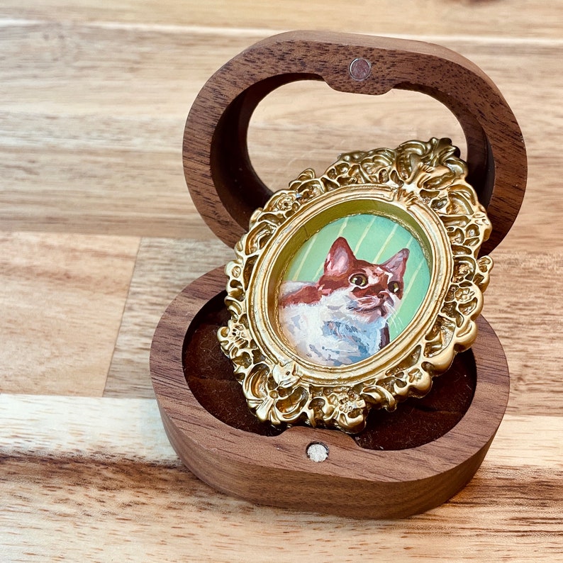 May include: A small, ornate gold frame with a painted portrait of a white and brown cat inside. The frame is sitting in a wooden box with a velvet lining.