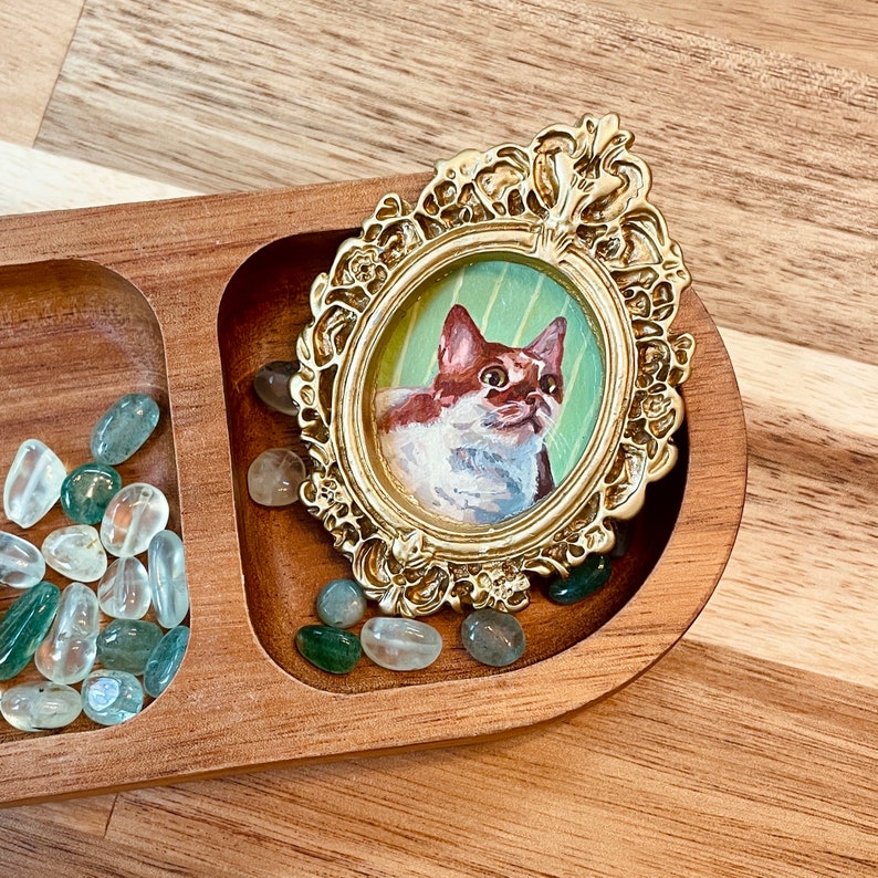 May include: A gold-toned ornate frame with a miniature painting of a white and brown cat with green eyes. The frame is sitting on a wooden tray with green and clear stones.