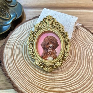 May include: A gold-colored ornate frame with a pink background and a painted portrait of a brown and white poodle.