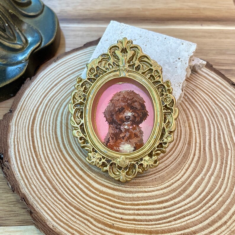 May include: A gold-colored ornate frame with a pink background and a painted portrait of a brown and white poodle.