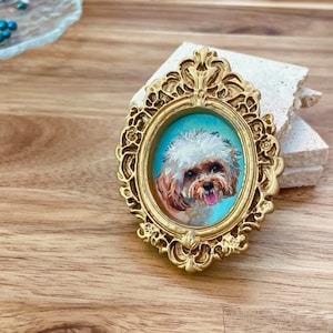 May include: A small, white and brown dog portrait in a gold ornate frame. The dog is looking directly at the viewer with its tongue sticking out.