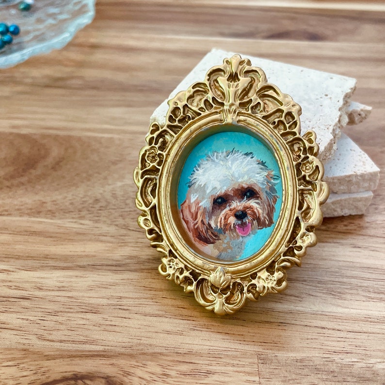 May include: A small, white and brown dog portrait in a gold ornate frame. The dog is looking directly at the viewer with its tongue sticking out.