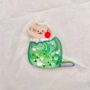 May include: A whimsical cat-shaped pin with a green, bubbly body and a light brown head. The cat has a red cherry on its head and a white frilly collar. The pin is set against a white, textured background.