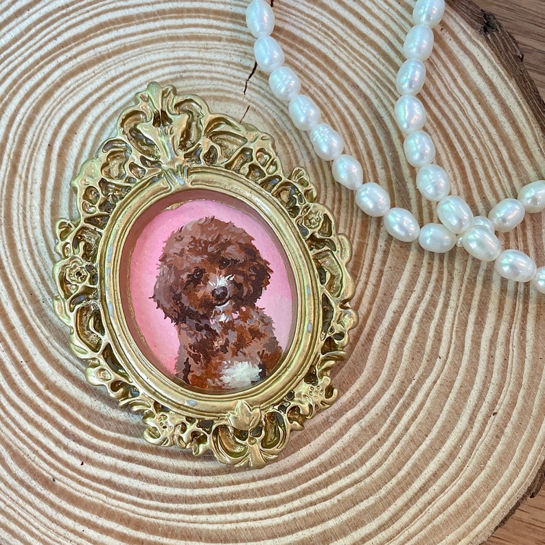 May include: A gold-toned ornate frame with a pink background and a painting of a brown and white poodle inside. The frame is sitting on a wooden surface.