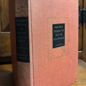 May include: A red hardcover book with the title "The Best Stories of Guy de Maupassant" by  Commins. The book is part of the Modern Library series.