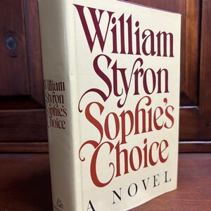 May include: A hardcover book titled "Sophie's Choice" by William Styron. The book's cover is off-white with the title and author's name in burgundy lettering. The book is a novel, as indicated on the cover.