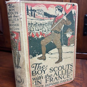 May include: A vintage book cover with a red, white, and blue color scheme. The cover features a boy scout holding a flag and marching with other soldiers in the background. The title of the book is "The Boy Scouts with the Allies in France" by Lieut. Howard Payson.