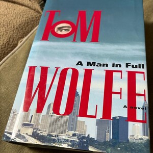 May include: A book cover with the title "A Man in Full" by Tom Wolfe. The cover features a red and white cityscape with a close-up of an eye peeking through the letter "O" in the author's name.