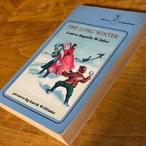 May include: A blue book titled "The Long Winter" by Laura Ingalls Wilder, with an illustration of children playing in the snow. The book is part of the Harper Trophy Book series.
