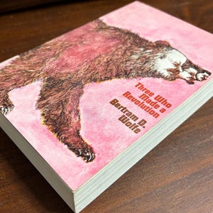 May include: A hardcover book titled "Three Who Made a Revolution" by Bertram D. Wolfe. The cover features a brown bear illustration on a pink background. The book's pages are white.