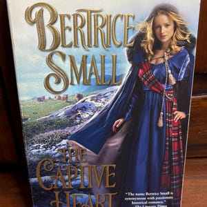 May include: A book cover for "The Captive Heart" by Bertrice Small. The cover features a woman in a blue dress and a red plaid scarf, set against a backdrop of a green landscape. The title and author's name are prominently displayed.