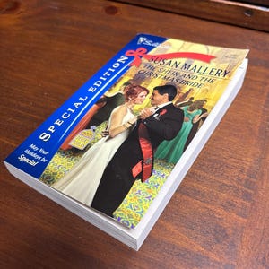 May include: A paperback book with a blue and white cover. The cover features a couple dancing in a ballroom. The title of the book is "The Sheik and the Christmas Bride" by Susan Mallery. The book is part of the "Desert Rogues" series.
