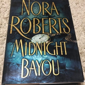 May include: A book cover with a dark blue background and gold lettering. The title is "Midnight Bayou" by Nora Roberts. A gold pocket watch with a white face is featured on the cover.