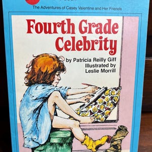 May include: A vintage paperback book titled "Fourth Grade Celebrity" by Patricia Reilly Giff, illustrated by Leslie Morrill. The cover features a young girl drawing, with text in red and blue. The book is a "Yearling Book" from Dell.