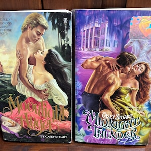 May include: Two paperback books with romantic cover art. One features a couple embracing with a sailboat in the background, titled "Moonlight Angel." The other, "Midnight Thunder," depicts a couple in front of a mansion. Both books are by Casey Stuart.
