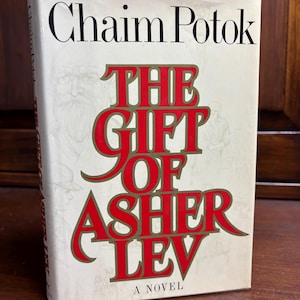 May include: A hardcover book titled "The Gift of Asher Lev" by Chaim Potok. The title is in large red letters with a gold outline. The author's name is in black letters at the top. The book has a cream-colored background.