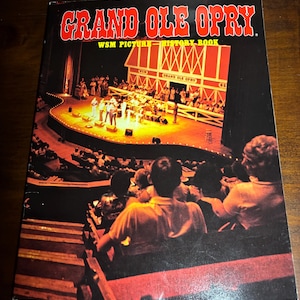 May include: A vintage book titled "Grand Ole Opry WSM Picture-History Book" with a photo of a stage with a band performing in front of a red barn with the words "Grand Ole Opry" on it. The photo shows a crowd of people watching the performance.
