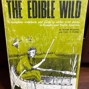 May include: A vintage cookbook titled "THE EDIBLE WILD" with a green cover. The cover features an illustration of a person in a canoe, with text describing the book as a guide to edible wild plants in Canada and North America.