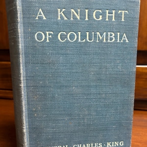 May include: A vintage book titled "A Knight of Columbia" by General Charles King. The book has a blue cover with white lettering. The book is standing upright, with the title and author clearly visible.